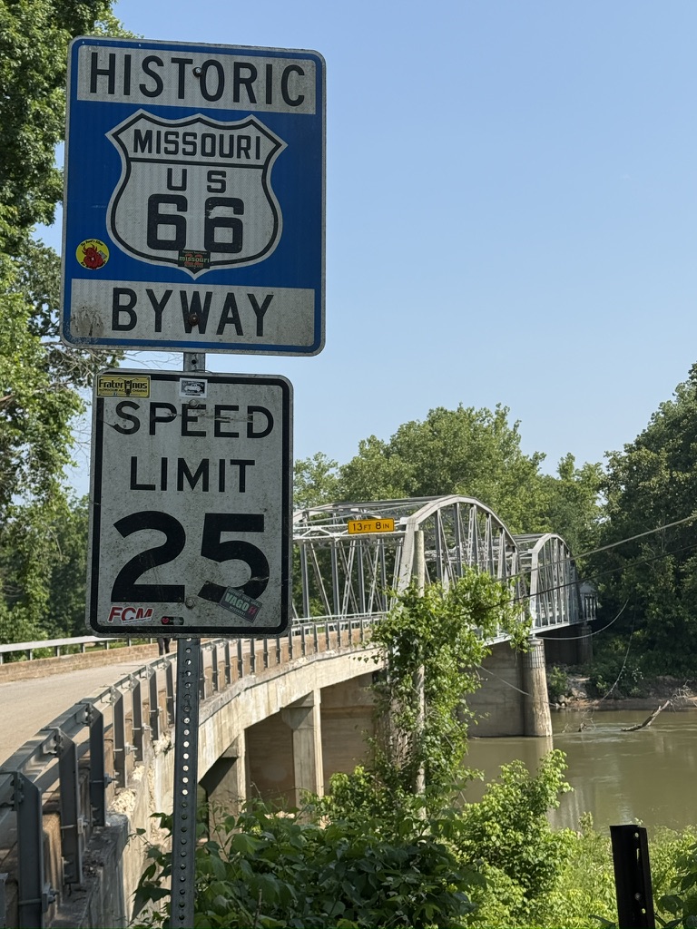 Schild „Historic Missouri US 66 Byway“ mit Tempo 25 vor einer Stahlbrücke über einen Fluss